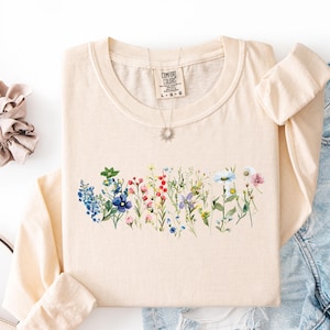 May include: Cream-colored long-sleeved shirt featuring a watercolor floral design across the chest. The design includes a variety of colorful wildflowers. A silver necklace with a sun pendant is visible. The shirt is folded, with a scrunchie and denim shorts nearby.