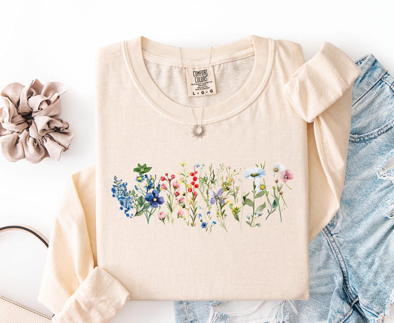 May include: Cream-colored long-sleeved shirt featuring a watercolor floral design across the chest. The design includes a variety of colorful wildflowers. A silver necklace with a sun pendant is visible. The shirt is folded, with a scrunchie and denim shorts nearby.