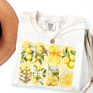 May include: Cream-colored t-shirt featuring a watercolor design of lemons, a bird, and a yellow bow. The design is divided into vertical panels. A gold necklace and a brown hat are also visible.
