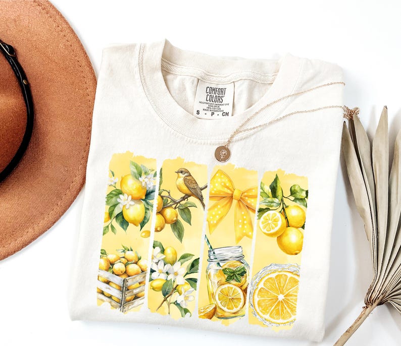 May include: Cream-colored t-shirt featuring a watercolor design of lemons, a bird, and a yellow bow. The design is divided into vertical panels. A gold necklace and a brown hat are also visible.