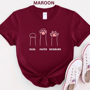 Puede incluir: Camiseta granate con un gr&aacute;fico blanco de patas de gato que representan piedra, papel y tijeras. El texto "ROCK PAPER SCISSORS" est&aacute; debajo. Zapatillas blancas con ribete rojo en la parte inferior derecha.