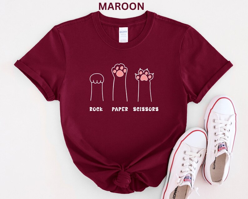 Puede incluir: Camiseta granate con un gr&aacute;fico blanco de patas de gato que representan piedra, papel y tijeras. El texto "ROCK PAPER SCISSORS" est&aacute; debajo. Zapatillas blancas con ribete rojo en la parte inferior derecha.