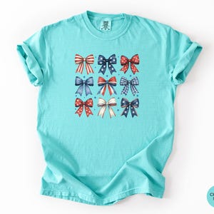 May include: A chalky mint short-sleeve t-shirt featuring a graphic print of nine decorative bows arranged in a grid. The bows are colored in red, white, and blue with various patterns including stripes, polka dots, and stars. The shirt has a crew neckline.