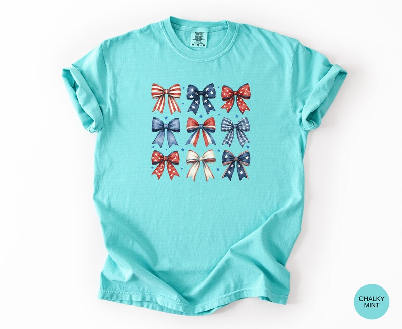 May include: A chalky mint short-sleeve t-shirt featuring a graphic print of nine decorative bows arranged in a grid. The bows are colored in red, white, and blue with various patterns including stripes, polka dots, and stars. The shirt has a crew neckline.