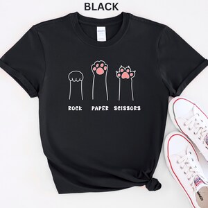 Puede incluir: Camiseta negra con un dise&ntilde;o divertido de patas de gato que representan piedra, papel y tijeras. El texto "ROCK PAPER SCISSORS" est&aacute; impreso debajo de las ilustraciones de las patas. Zapatillas blancas con detalles rojos tambi&eacute;n son visibles.
