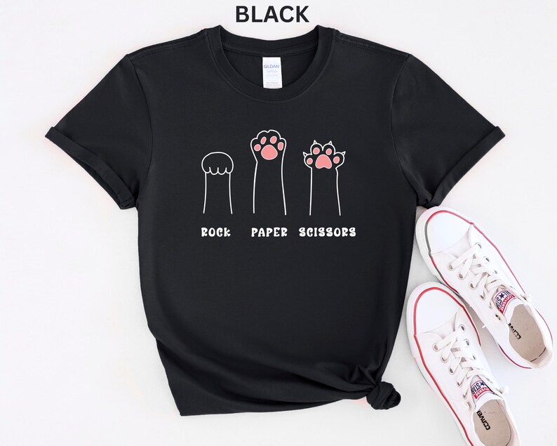 Puede incluir: Camiseta negra con un dise&ntilde;o divertido de patas de gato que representan piedra, papel y tijeras. El texto "ROCK PAPER SCISSORS" est&aacute; impreso debajo de las ilustraciones de las patas. Zapatillas blancas con detalles rojos tambi&eacute;n son visibles.