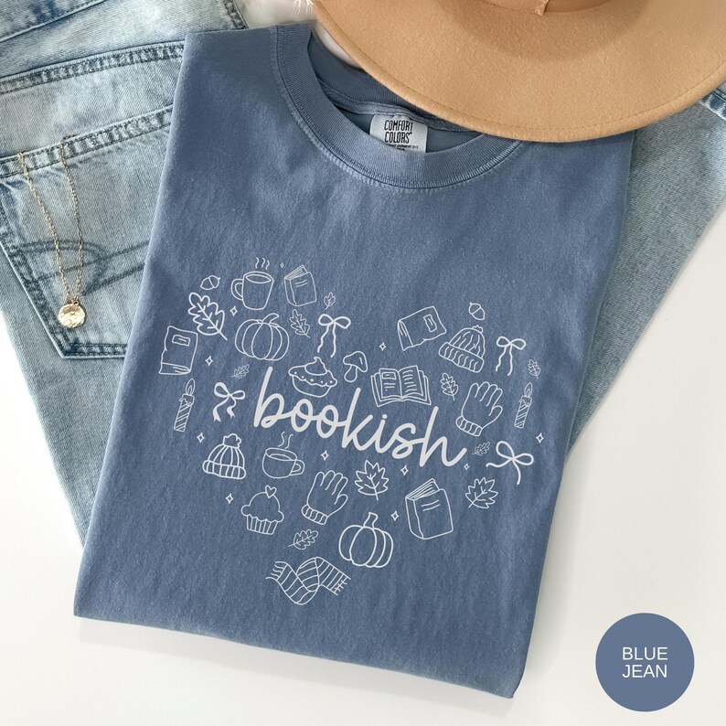 May include: A blue t-shirt featuring a white graphic print of the word bookish surrounded by various line art icons including pumpkins, books, mugs, hats, and leaves arranged in a heart shape. The shirt has a Comfort Colors brand label.