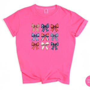 May include: A bright pink short-sleeved t-shirt featuring a graphic print of nine decorative bows arranged in a three-by-three grid. The bows display various patterns including red and white stripes, blue with white stars, and red with white polka dots.