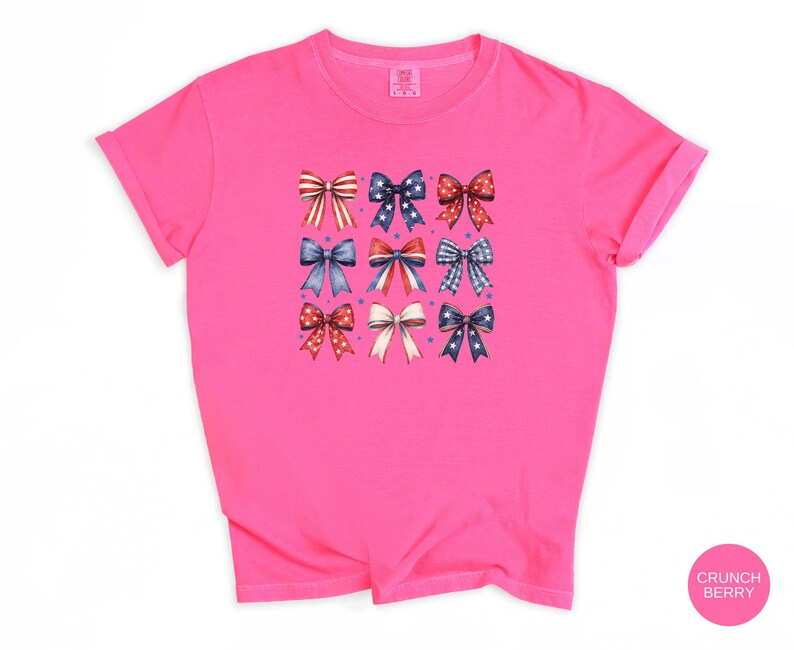 May include: A bright pink short-sleeved t-shirt featuring a graphic print of nine decorative bows arranged in a three-by-three grid. The bows display various patterns including red and white stripes, blue with white stars, and red with white polka dots.