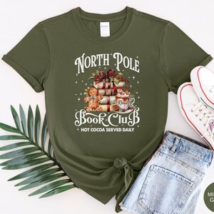 May include: An olive green short-sleeve t-shirt featuring a graphic print of a stack of books, a gingerbread man, and a mug of hot cocoa. The text reads North Pole Book Club and Hot Cocoa Served Daily. The design includes holly and stars.