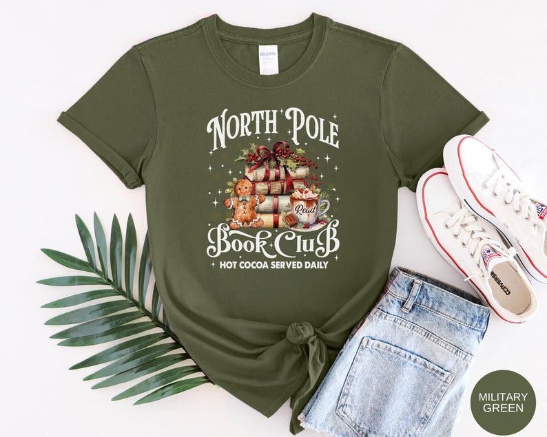 May include: An olive green short-sleeve t-shirt featuring a graphic print of a stack of books, a gingerbread man, and a mug of hot cocoa. The text reads North Pole Book Club and Hot Cocoa Served Daily. The design includes holly and stars.