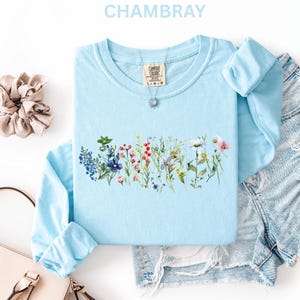 May include: Light blue long-sleeve shirt with a floral design across the chest. The shirt has a crew neck and is made of a soft material. The word "CHAMBRAY" is at the top of the image. A scrunchie, purse, and denim shorts are also in the image.