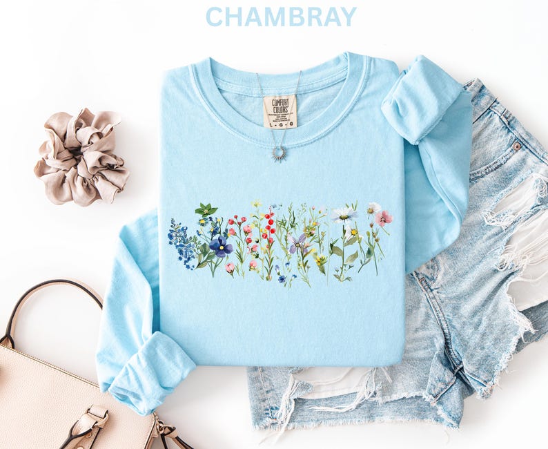 May include: Light blue long-sleeve shirt with a floral design across the chest. The shirt has a crew neck and is made of a soft material. The word "CHAMBRAY" is at the top of the image. A scrunchie, purse, and denim shorts are also in the image.