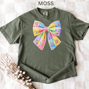 May include: A moss green t-shirt with a large, colourful bow design. The bow is made of patchwork fabric with floral and polka dot patterns in pink, yellow, blue, and green. The word "MOSS" is printed at the top.