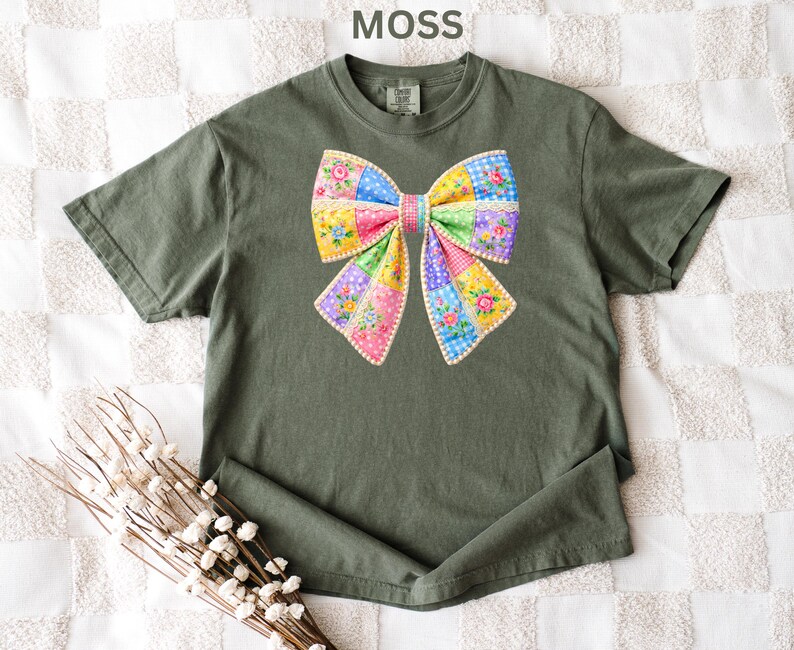 May include: A moss green t-shirt with a large, colourful bow design. The bow is made of patchwork fabric with floral and polka dot patterns in pink, yellow, blue, and green. The word "MOSS" is printed at the top.