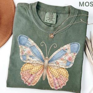 May include: Moss green t-shirt with a patchwork butterfly design. The butterfly wings feature denim, floral, and plaid patterns in blue, pink, yellow, and cream. A delicate gold necklace with a pendant rests on the shirt. The shirt has a Comfort Colors tag.