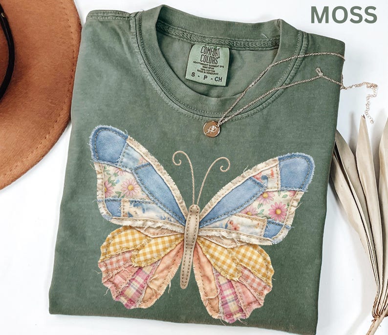 May include: Moss green t-shirt with a patchwork butterfly design. The butterfly wings feature denim, floral, and plaid patterns in blue, pink, yellow, and cream. A delicate gold necklace with a pendant rests on the shirt. The shirt has a Comfort Colors tag.