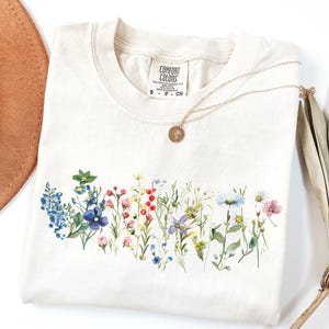 May include: Cream-colored t-shirt with a watercolor floral design across the chest. The design features a variety of colorful wildflowers. A gold necklace with a pendant rests on the shirt. The shirt has a Comfort Colors tag.