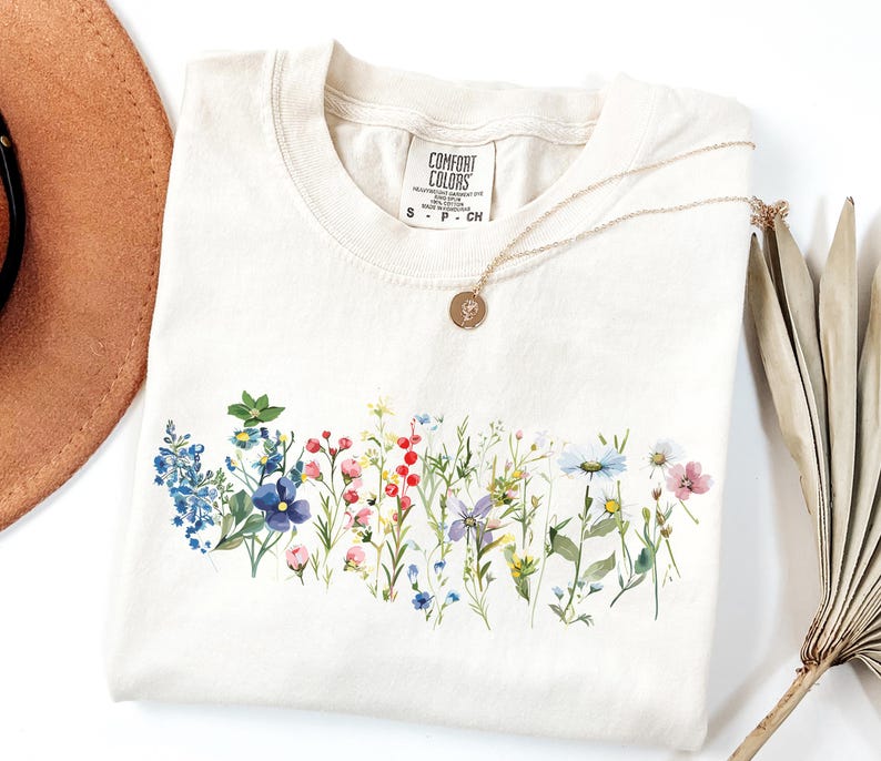 May include: Cream-colored t-shirt with a watercolor floral design across the chest. The design features a variety of colorful wildflowers. A gold necklace with a pendant rests on the shirt. The shirt has a Comfort Colors tag.