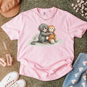 Puede incluir: Una camiseta rosa claro con un gr&aacute;fico de dos monos de dibujos animados abraz&aacute;ndose. Un mono es gris y el otro es marr&oacute;n. La camiseta tiene cuello redondo y mangas cortas. La camiseta est&aacute; hecha de un material suave.
