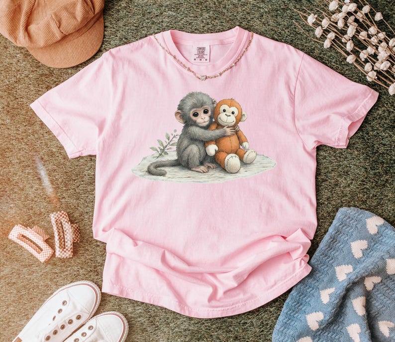 Puede incluir: Una camiseta rosa claro con un gr&aacute;fico de dos monos de dibujos animados abraz&aacute;ndose. Un mono es gris y el otro es marr&oacute;n. La camiseta tiene cuello redondo y mangas cortas. La camiseta est&aacute; hecha de un material suave.