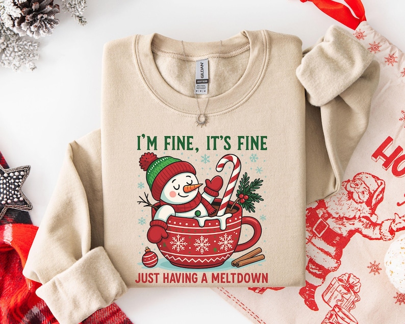 Pu&ograve; includere: Una felpa beige a girocollo con la grafica di un pupazzo di neve seduto in una tazza rossa con motivi a fiocchi di neve bianchi. Il testo recita I'M FINE, IT'S FINE e JUST HAVING A MELTDOWN. Il pupazzo di neve indossa un cappello.