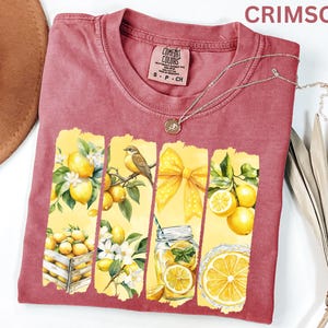 May include: Crimson Comfort Colors t-shirt with a lemon-themed graphic. The design features yellow panels with lemons, a bird, a bow, and a jar of lemonade. A silver necklace with a pendant is draped across the shirt.