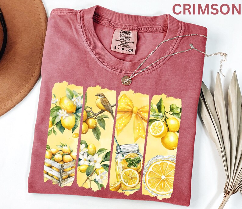 May include: Crimson Comfort Colors t-shirt with a lemon-themed graphic. The design features yellow panels with lemons, a bird, a bow, and a jar of lemonade. A silver necklace with a pendant is draped across the shirt.