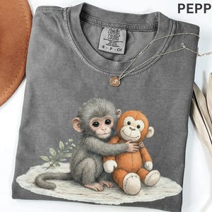 Puede incluir: Una camiseta gris Comfort Colors con un estampado de un mono gris abrazando a un mono de peluche naranja. La camiseta tiene cuello redondo y mangas cortas. Un collar plateado con un peque&ntilde;o colgante est&aacute; sobre la camiseta.