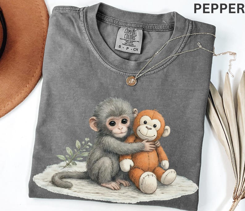 Puede incluir: Una camiseta gris Comfort Colors con un estampado de un mono gris abrazando a un mono de peluche naranja. La camiseta tiene cuello redondo y mangas cortas. Un collar plateado con un peque&ntilde;o colgante est&aacute; sobre la camiseta.