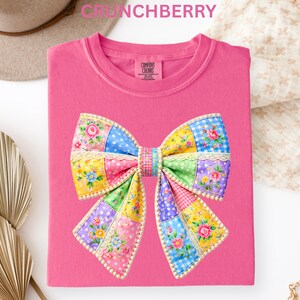 May include: A pink Comfort Colors t-shirt featuring a large, colorful bow design. The bow is made of patchwork squares in various colors and patterns, including floral prints, polka dots, and gingham. The word "CRUNCHBERRY" is at the top.