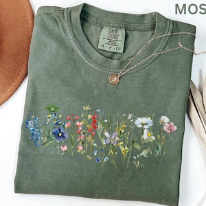 May include: A moss green Comfort Colors t-shirt with a floral design across the front. The shirt has a crew neck and short sleeves. A silver necklace with a small pendant rests on the shirt. The word "MOSS" is in the upper right corner.