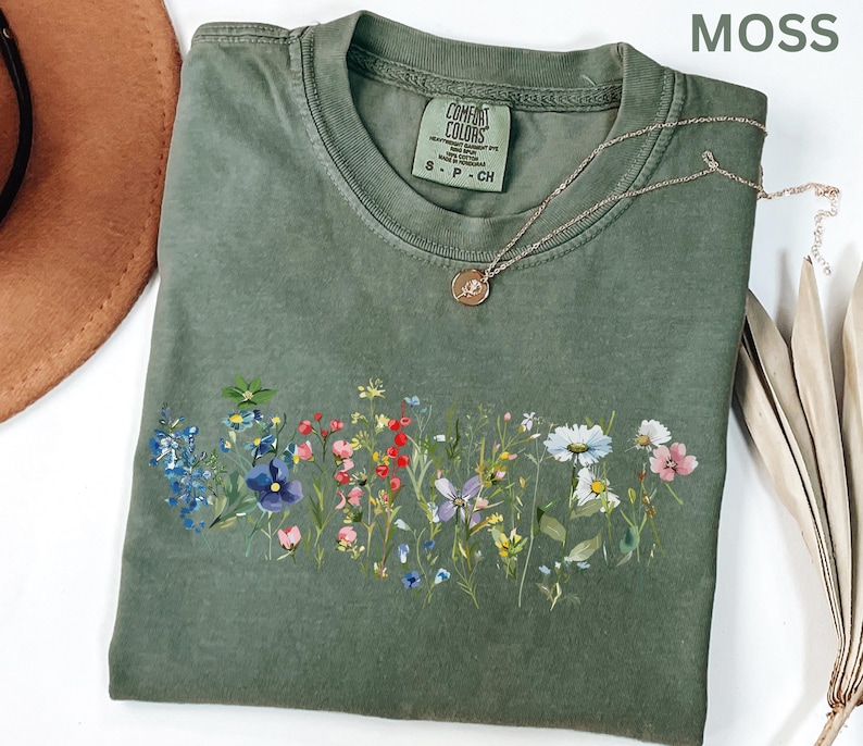 May include: A moss green Comfort Colors t-shirt with a floral design across the front. The shirt has a crew neck and short sleeves. A silver necklace with a small pendant rests on the shirt. The word "MOSS" is in the upper right corner.