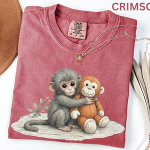 Puede incluir: Camiseta de color carmes&iacute; con un gr&aacute;fico de dos monos abraz&aacute;ndose. Un mono es gris y el otro naranja. La camiseta tiene cuello redondo y mangas cortas. Un collar plateado con un colgante est&aacute; drapeado sobre la camiseta.