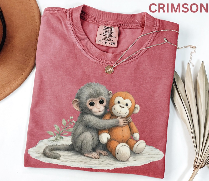 Puede incluir: Camiseta de color carmes&iacute; con un gr&aacute;fico de dos monos abraz&aacute;ndose. Un mono es gris y el otro naranja. La camiseta tiene cuello redondo y mangas cortas. Un collar plateado con un colgante est&aacute; drapeado sobre la camiseta.