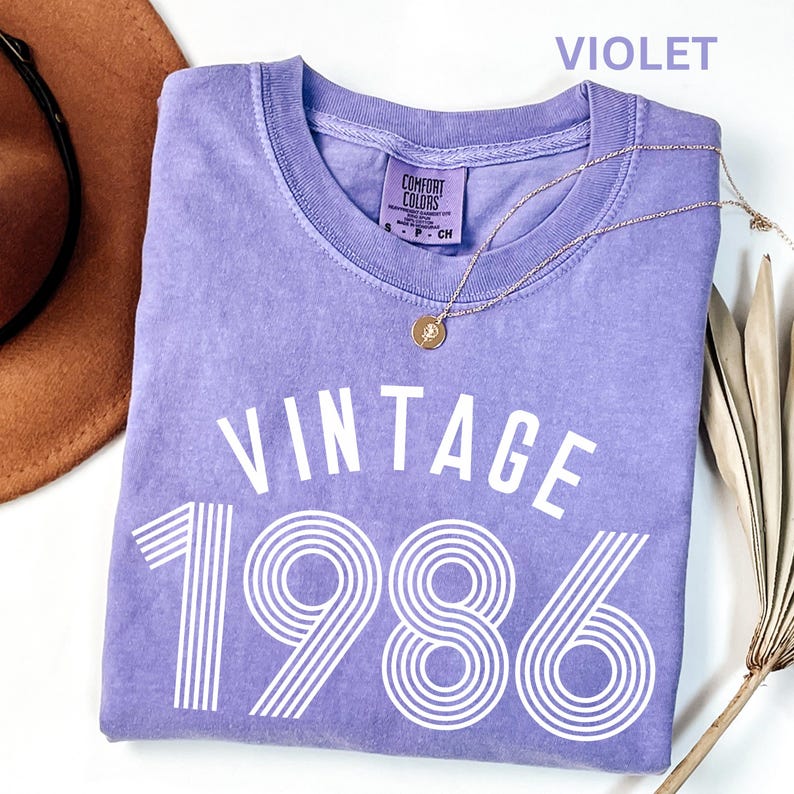 May include: A violet-colored t-shirt with the text "VINTAGE 1986" in white. The shirt has a crew neck and a small tag that says "COMFORT COLORS". A gold necklace with a pendant is draped across the shirt. A brown hat is visible in the upper left corner.