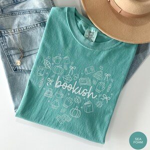 May include: A seafoam green t-shirt featuring a white graphic print of the word bookish surrounded by line art illustrations of pumpkins, books, mugs, leaves, bows, mittens, and hats. The shirt has a Comfort Colors label at the inner neckline.