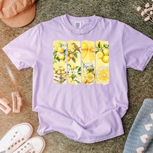 May include: A light purple t-shirt featuring a watercolor design of lemons, a bird, and a yellow bow. The design is arranged in vertical rectangular panels. The shirt is surrounded by a hat, shoes, and a blanket.