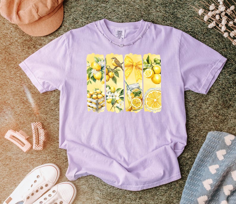 May include: A light purple t-shirt featuring a watercolor design of lemons, a bird, and a yellow bow. The design is arranged in vertical rectangular panels. The shirt is surrounded by a hat, shoes, and a blanket.