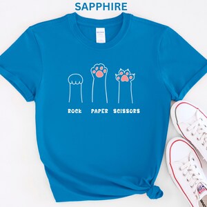 Puede incluir: Camiseta azul zafiro con un gr&aacute;fico blanco de tres patas de gato que representan piedra, papel y tijeras. Las patas tienen almohadillas rosas. El texto blanco debajo de las patas dice "ROCK PAPER SCISSORS". Zapatillas blancas visibles en la esquina inferior derecha.
