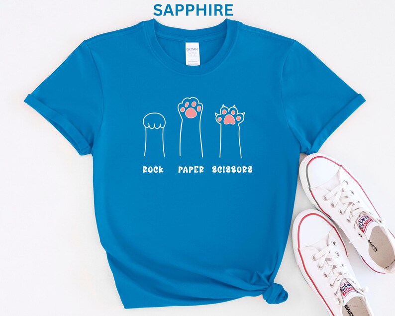 Puede incluir: Camiseta azul zafiro con un gr&aacute;fico blanco de tres patas de gato que representan piedra, papel y tijeras. Las patas tienen almohadillas rosas. El texto blanco debajo de las patas dice "ROCK PAPER SCISSORS". Zapatillas blancas visibles en la esquina inferior derecha.