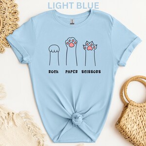 Puede incluir: Camiseta azul claro con un dise&ntilde;o l&uacute;dico de "Piedra, Papel, Tijeras" utilizando ilustraciones de patas de gato. El texto "LIGHT BLUE" est&aacute; en la parte superior. La camiseta est&aacute; anudada en la parte inferior.