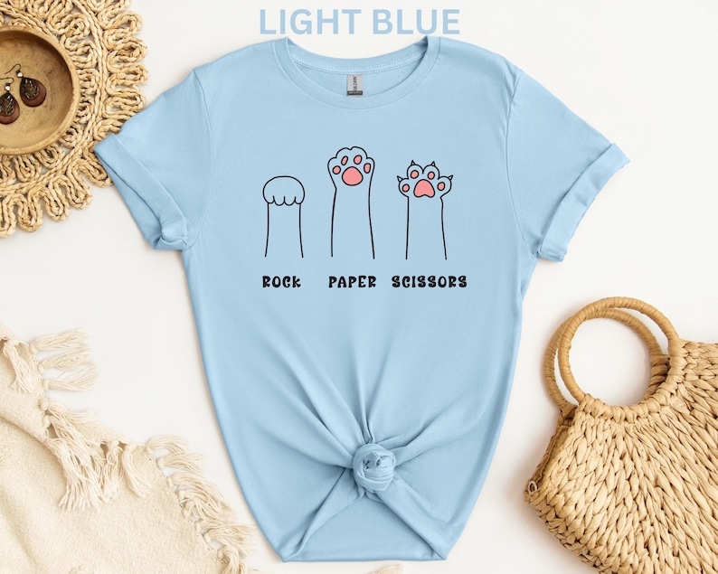 Puede incluir: Camiseta azul claro con un dise&ntilde;o l&uacute;dico de "Piedra, Papel, Tijeras" utilizando ilustraciones de patas de gato. El texto "LIGHT BLUE" est&aacute; en la parte superior. La camiseta est&aacute; anudada en la parte inferior.