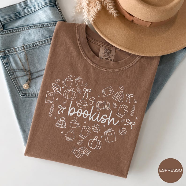 May include: A brown t-shirt featuring a white graphic of the word bookish surrounded by small line art icons including pumpkins, books, mugs, leaves, and winter accessories. The shirt is layered with blue denim jeans and a tan wide-brimmed hat.