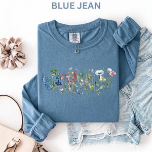 May include: A blue jean long-sleeve shirt with a floral design. The shirt has a crew neck and is paired with denim shorts, a light pink purse, and a beige scrunchie. The shirt has the text "BLUE JEAN" at the top.