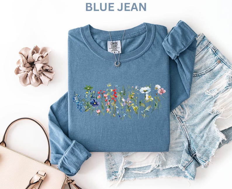 May include: A blue jean long-sleeve shirt with a floral design. The shirt has a crew neck and is paired with denim shorts, a light pink purse, and a beige scrunchie. The shirt has the text "BLUE JEAN" at the top.
