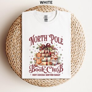 A white t-shirt with a graphic design featuring a gingerbread man, a stack of books, and a cup of hot cocoa, along with the text "North Pole Book Club" and "Hot Cocoa Served Daily".