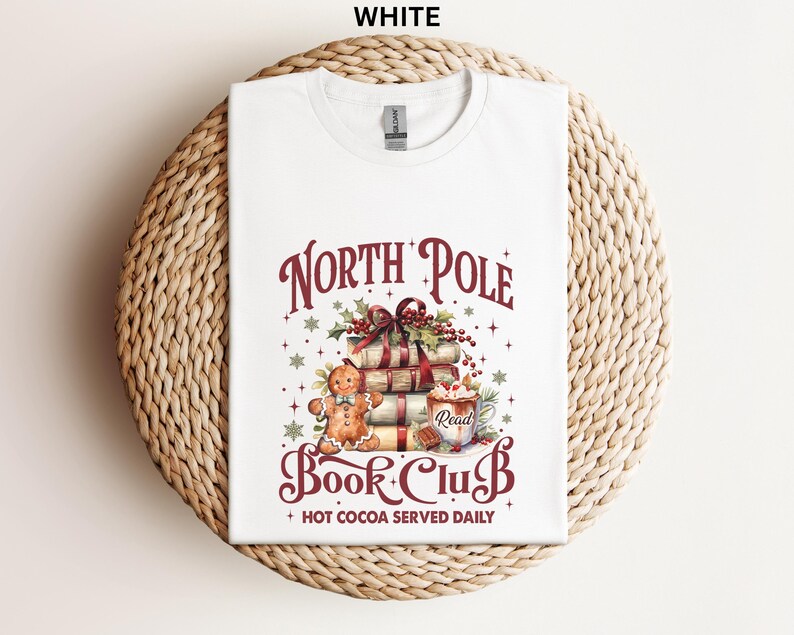 A white t-shirt with a graphic design featuring a gingerbread man, a stack of books, and a cup of hot cocoa, along with the text "North Pole Book Club" and "Hot Cocoa Served Daily".