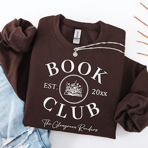 May include: A dark chocolate brown crewneck sweatshirt featuring white screen-printed text that reads BOOK CLUB, EST. 20xx, and The Glengreen Readers. The design includes a circular graphic of an open book with flowers emerging from the pages.