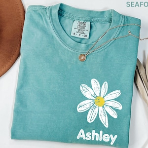 May include: Seafoam green t-shirt with a daisy graphic and the name "Ashley" printed in white. The shirt has a crew neck and short sleeves. A gold necklace with a pendant is draped across the shirt.